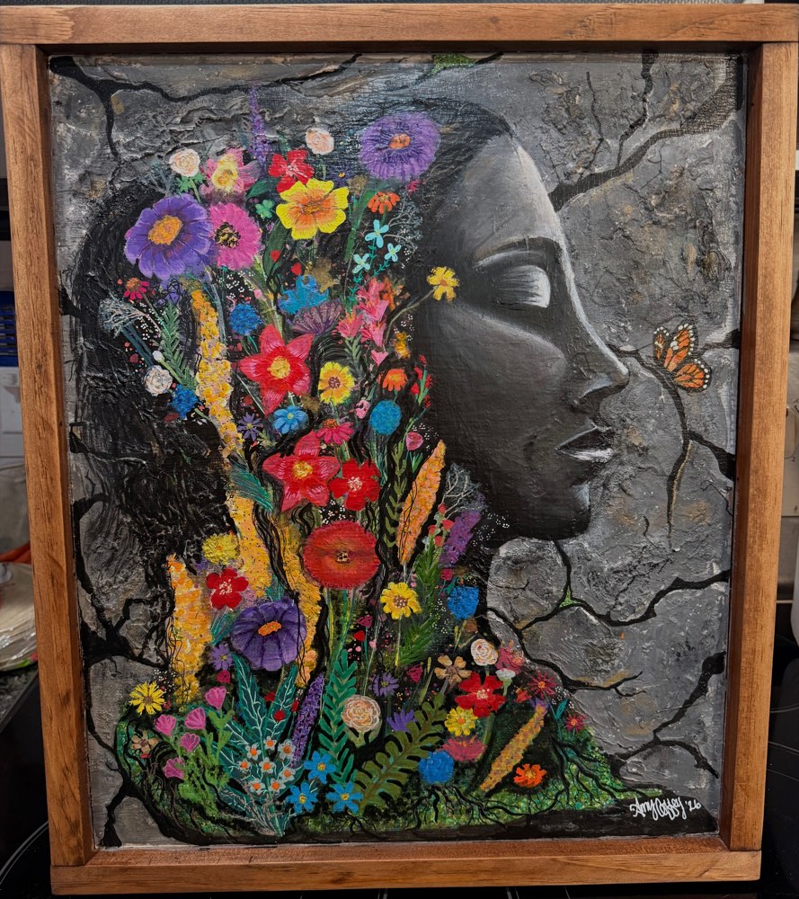 She Blooms Anyway by Amy Coffey — a woman's profile in grey against cracked ground, her hair made of dense painted wildflowers, in a reclaimed-wood frame.