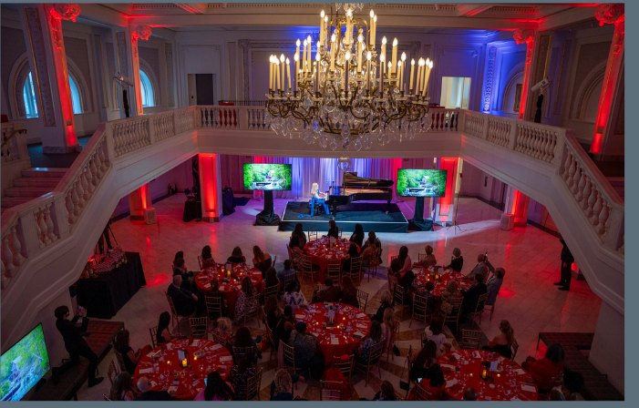 The ballroom set for dinner under a chandelier at a past Power Up opening night, with a pianist on stage